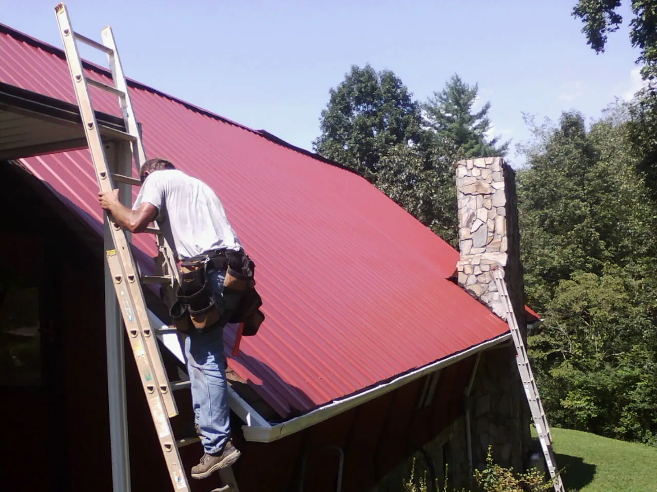 Roofer on a ladder beside a red metal roof and stone chimney — Soffit & Fascia Repair by Roofing Brothers in Gibbsboro
